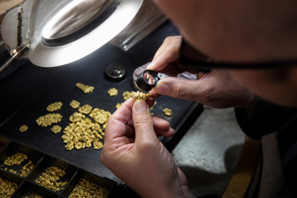 Jeweler's hands holding a loupe examining raw gold flakes under bright magnification lamp, close-up detail showing gold texture and authenticity inspection