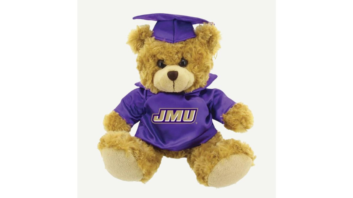 A flat lay of JMU graduation gifts arranged on a purple background: a gold diploma frame holding a 5x7 photo, a small plush bear wearing a graduation cap, a folded royal blue JMU t-shirt, and a small yard sign with metal stakes