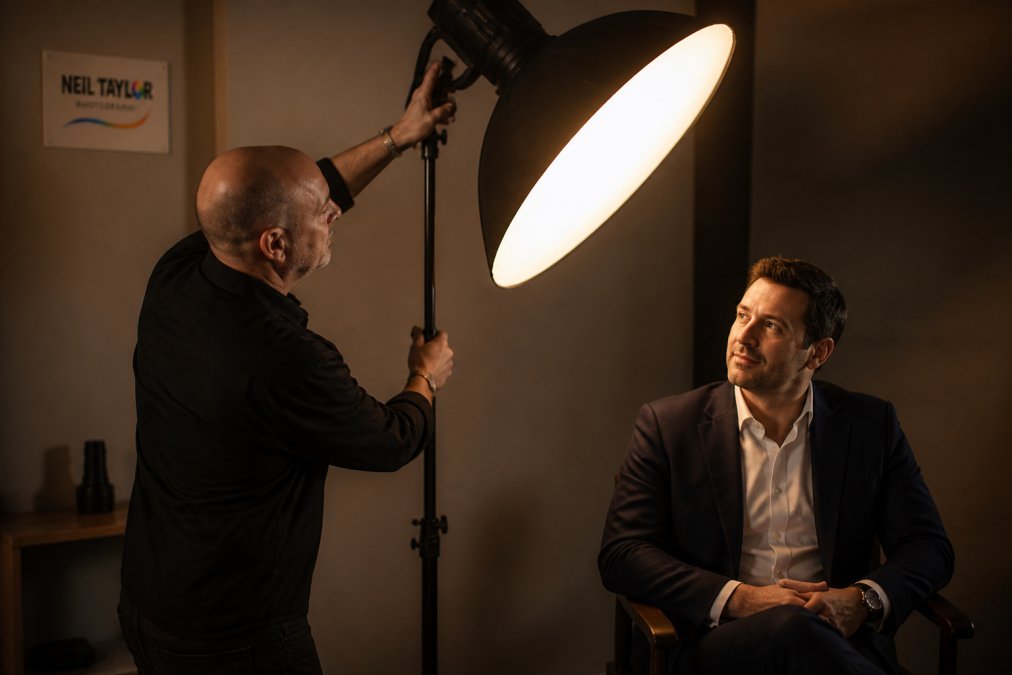 Professional photographer adjusting studio lighting equipment while model sits relaxed in chair, soft directional light illuminating face, neutral background