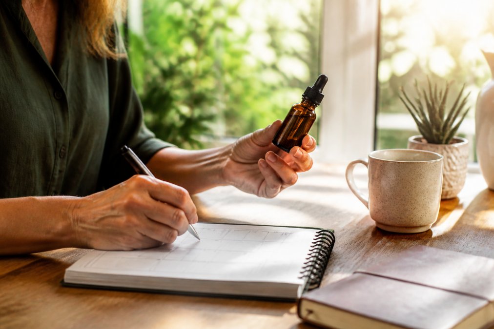 Person holding a CBD oil tincture bottle with dropper in one hand while writing dosage notes on a calendar on a wooden desk, with natural morning sunlight streaming through a window