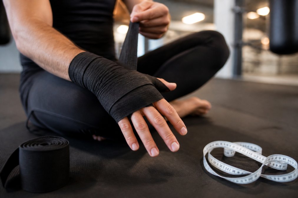 Close-up van een persoon die zijn hand inpakt met boksbandages op een gymmat, met een meetlint naast de hand liggend op de mat, zachte sportschoolverlichting op de achtergrond