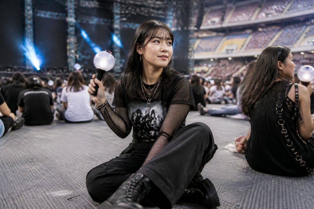 Young fan wearing a custom graphic tee layered with a sheer mesh top, oversized cargo pants, and platform shoes at a concert venue, holding an ARMY bomb with stage lights in background