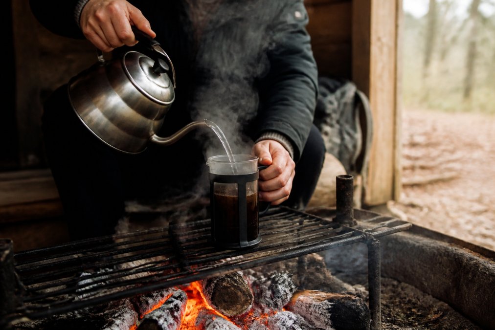 Person brygger kaffe over åbent bål i shelter, hælder varmt vand ind i stempelkande med damp stigende, glødende kulstykker synlige under en grillrist, naturligt dagslys