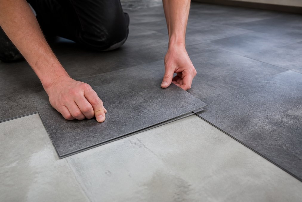 To hænder monterer kliksystem PVC-fliser på betonundergrund, viser hvordan fliserne presses sammen til de klikker på plads, med flere fliser allerede lagt i baggrunden