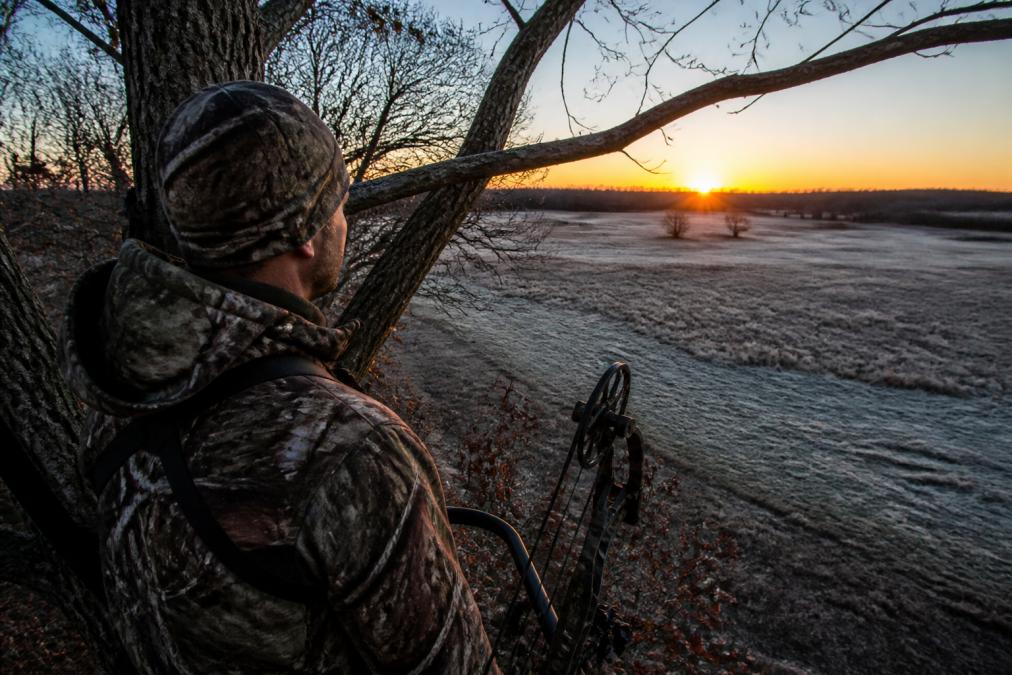 A hunter in camouflage standing in a tree stand at dawn, looking out over an open field with frost on the grass, showing the solitude and mixed emotions of early morning hunting