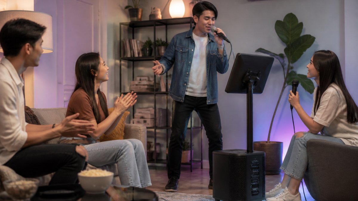 Friends singing with a wireless microphone near an Ikarao karaoke machine in a bright, colorful living room