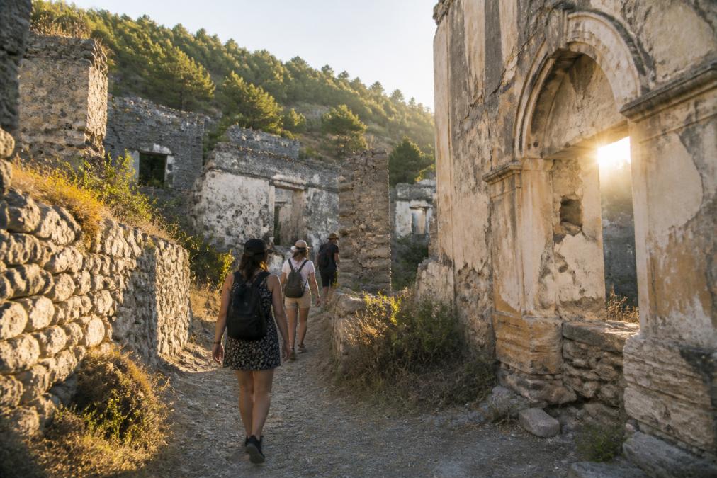 Ziyaretçilerin Kayaköy'ün tarihi taş sokakları arasında yürüdüğü, kilise kalıntılarını ve eski evleri incelediği sahne, güneşin duvarlar arasından filtre edildiği an