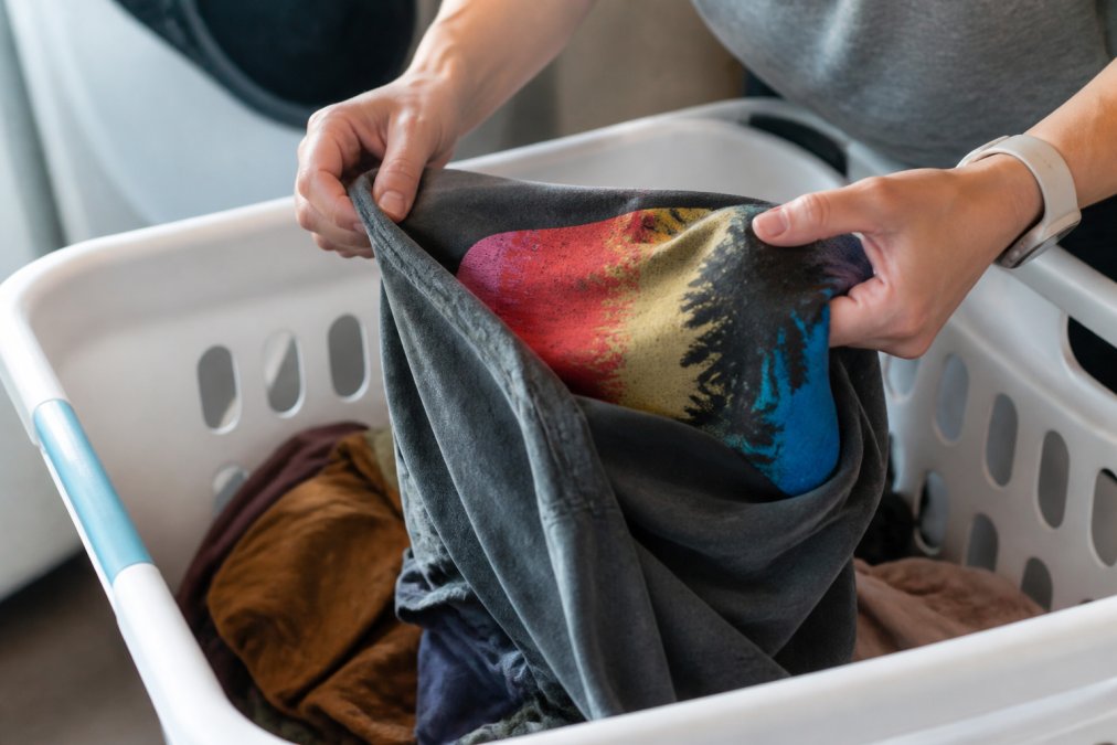 Close-up of hands carefully turning a colorful graphic t-shirt inside out over a laundry basket, showing the vibrant screen-printed design on the reverse side and the care being taken