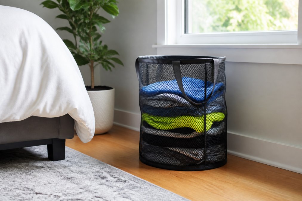 Open mesh laundry basket filled with folded gym clothes positioned in a bedroom corner with natural light and clear airflow around the basket, showing proper ventilation and storage setup
