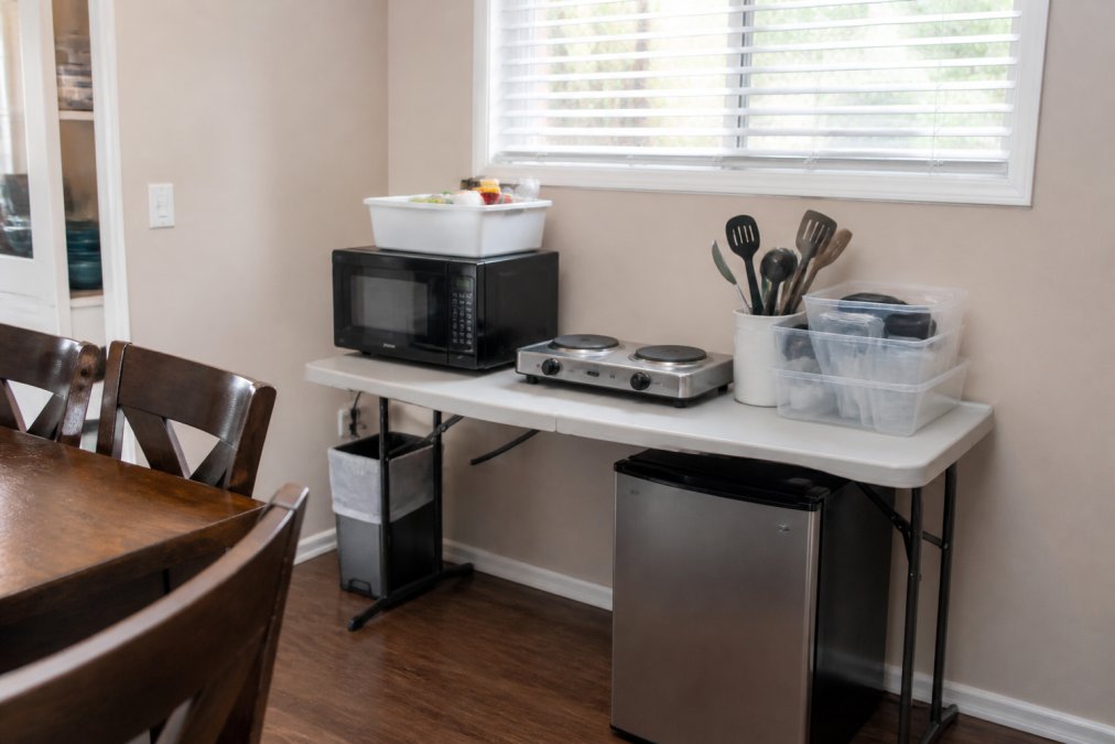 Makeshift kitchen setup in a dining room corner with a microwave, hot plate, and compact refrigerator on a folding table, basic dishes and utensils organized in clear containers nearby, natural window light