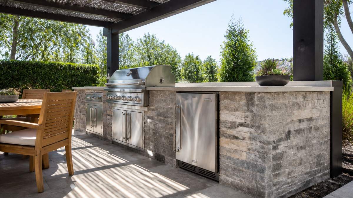 Professional outdoor kitchen with refrigerator installed under a pergola with shade cloth overhead, showing clear ventilation space around the unit with condenser coils exposed to airflow and natural shade pattern on surrounding patio