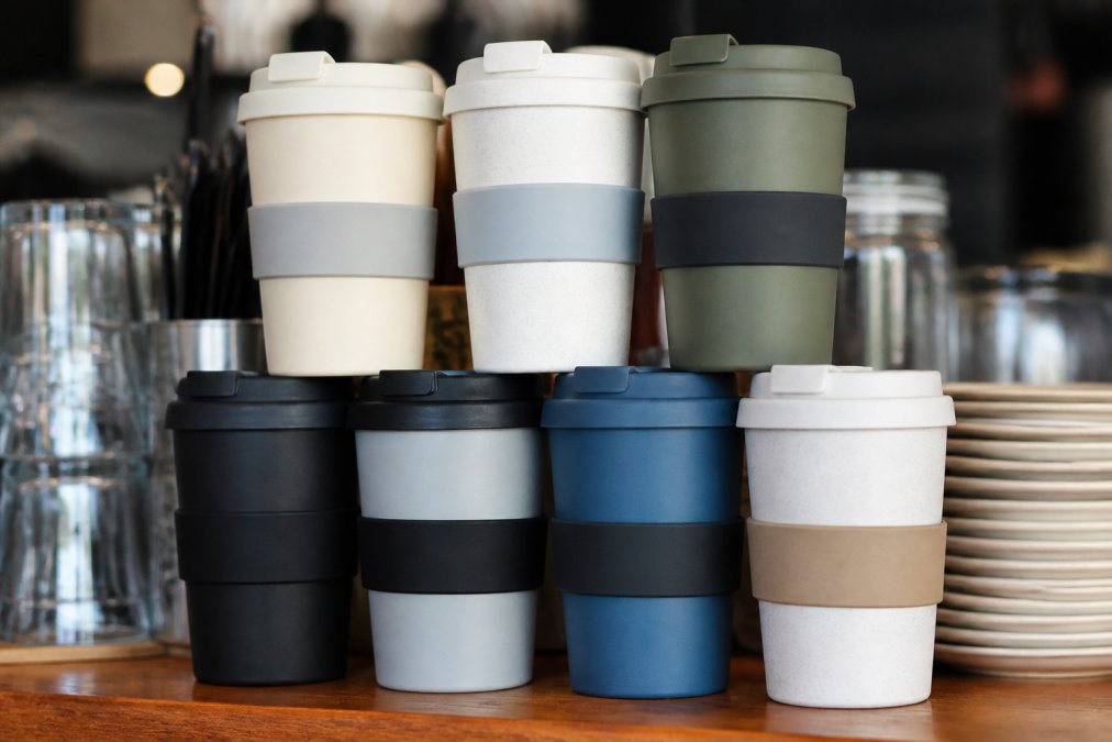 Close-up of stacked branded reusable coffee cups with custom logos and thermal insulation sleeves displayed on a cafe counter, showing leak-proof lids in natural daylight