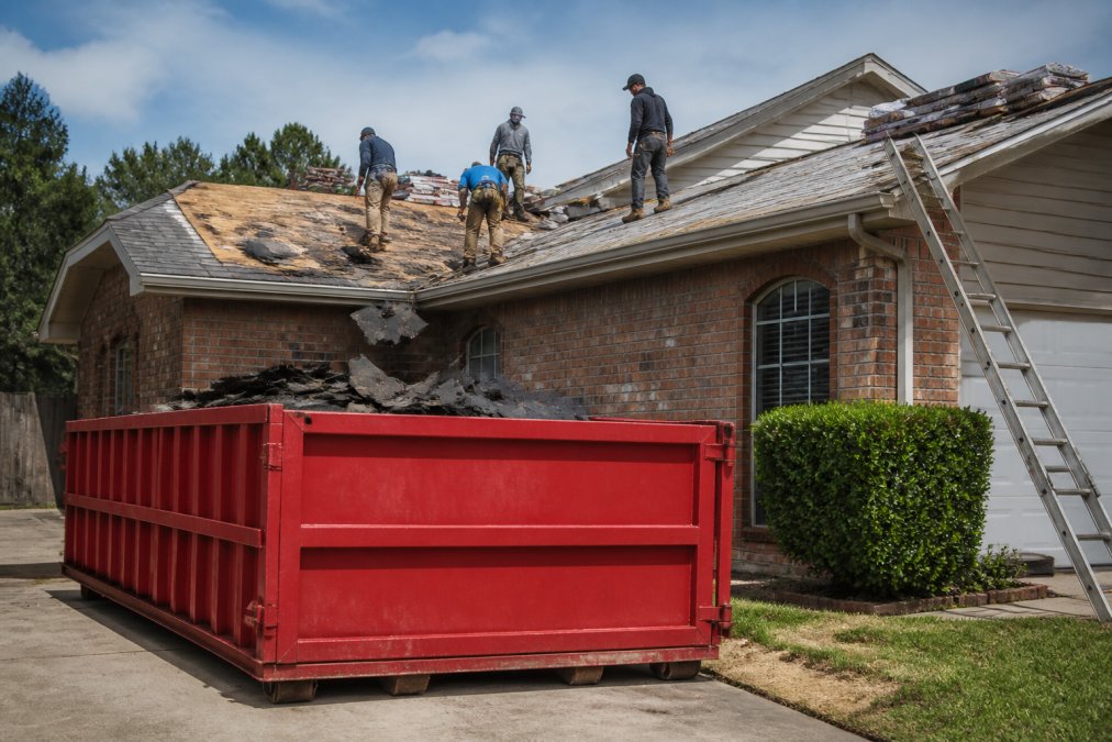 Professional roofing crew actively removing old shingles and materials from a residential roof, with a roll-off dumpster positioned on the driveway below to catch debris falling from the roof