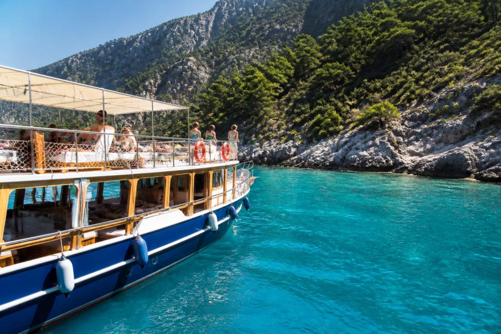 Ölüdeniz'in turkuaz sularında süzülen bir tekne, güvertede güneşlenen ve manzarayı izleyen tatilciler, arka planda yemyeşil koylar ve dağlar