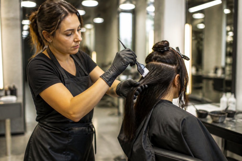 A professional stylist in a modern salon sectioning a client's hair and applying a keratin treatment with a brush, wearing gloves, under bright overhead lighting