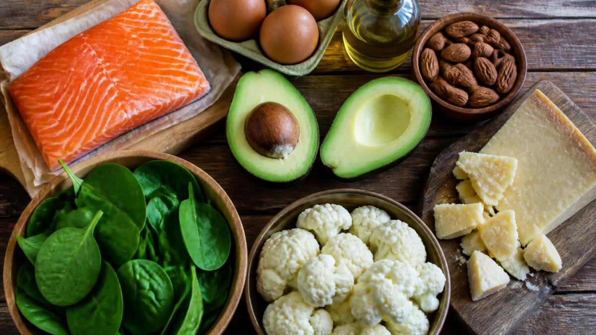 Overhead flat-lay of keto-friendly foods arranged on a wooden table: fresh salmon fillet, eggs, avocado halves, handful of almonds, fresh spinach leaves, raw cauliflower florets, olive oil in a small glass bottle, and blocks of aged cheese