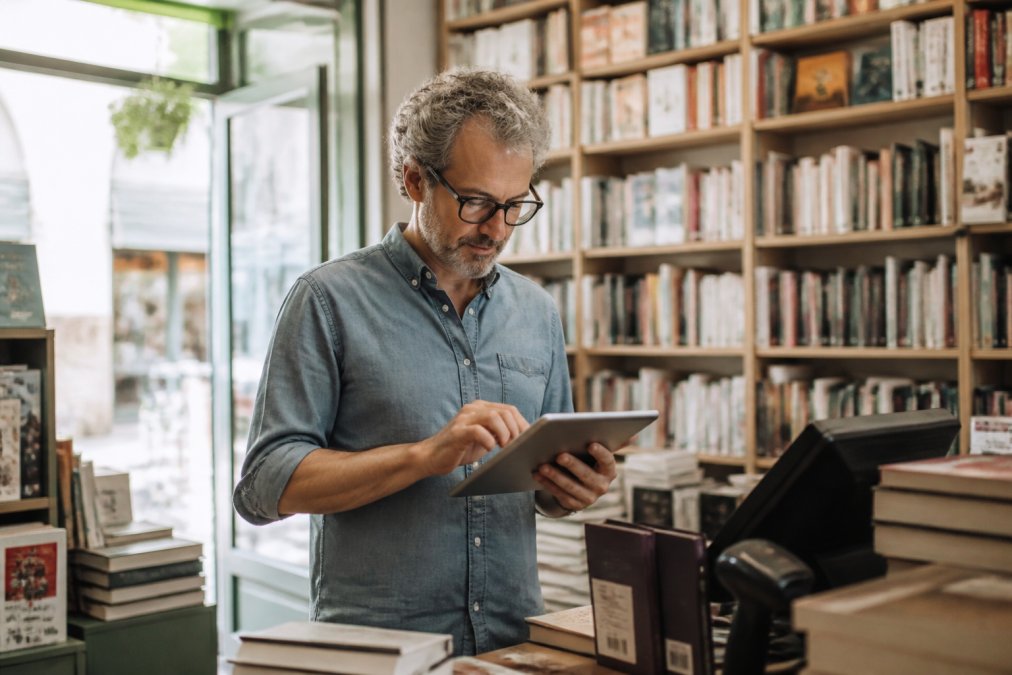 Il titolare di una libreria indipendente consulta i dati di vendita su un tablet al bancone della cassa, circondato da scaffali pieni di libri, con luce naturale che filtra dalla vetrina del negozio