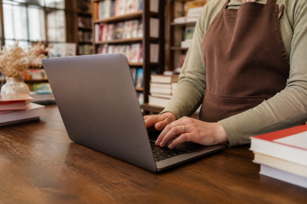 Primo piano di mani che utilizzano un laptop che mostra un pannello di controllo cloud con metriche di inventario e vendite, su una scrivania in una piccola libreria indipendente con scaffali di libri sullo sfondo