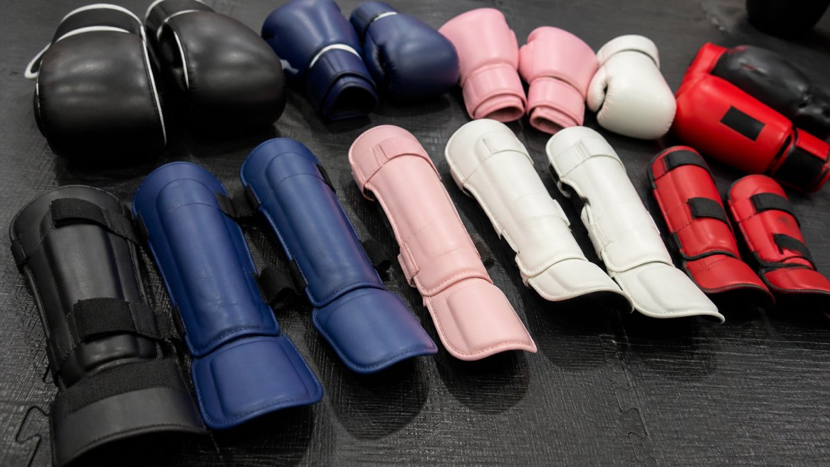 Close-up van verschillende bokshandschoenen en scheenbeschermers in diverse kleuren en maten uitgestald op een trainingsmat, met duidelijk zichtbare details van de materialen en sluitingen
