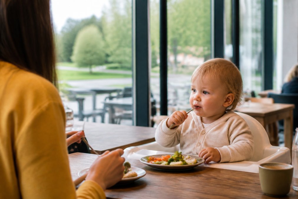 Toddler in a high chair at a cafe table enjoying a simple meal, with a parent nearby and green park scenery visible through bright windows in the background