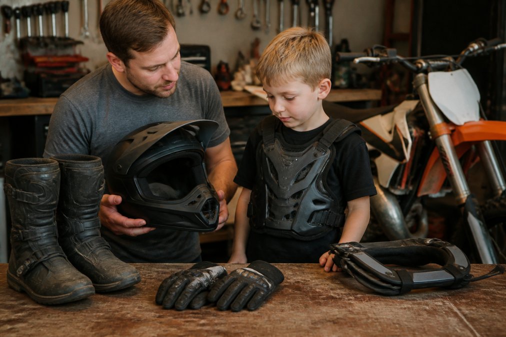 Parent and young child examining dirt bike protective gear together on a workbench, checking helmet fit and chest protector placement, with boots and gloves laid out for inspection