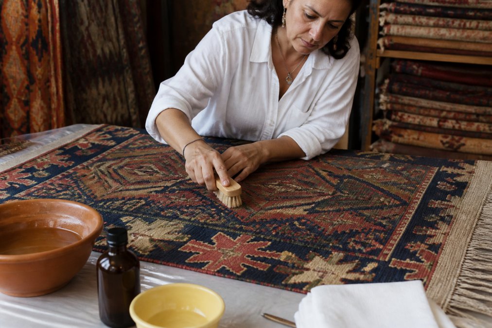Person carefully cleaning an antique kilim rug with soft-bristled brush and natural cleaning solution, demonstrating gentle maintenance technique in well-lit workspace with the rug laid flat