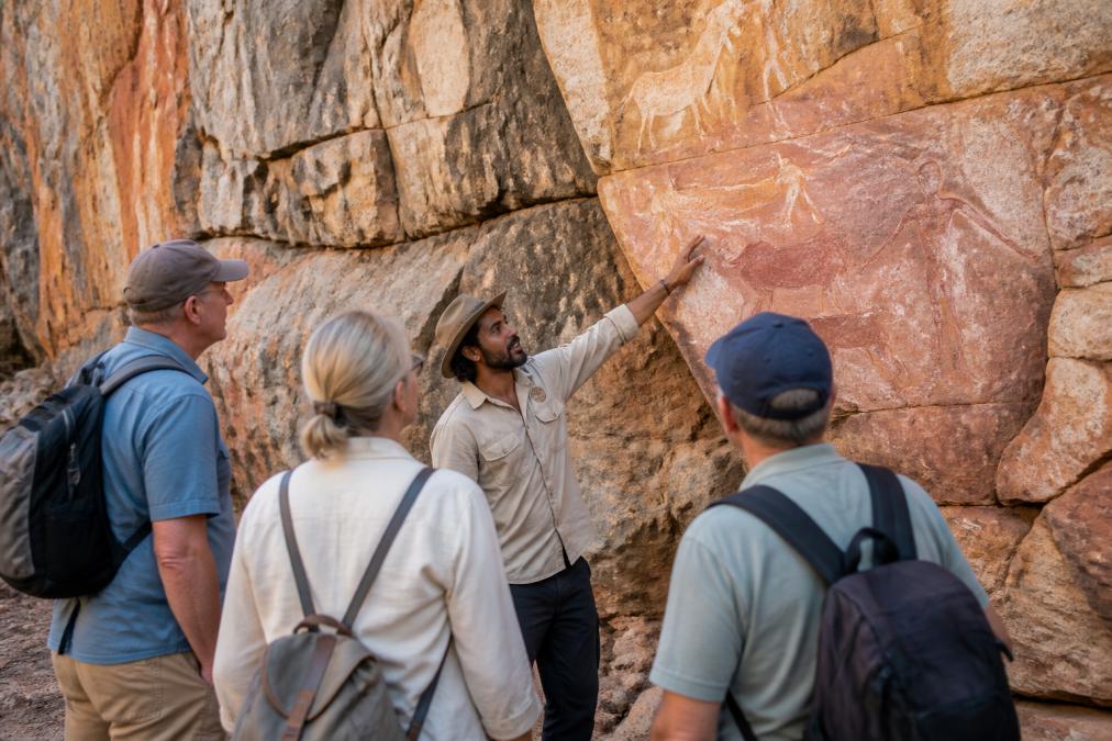 Local Aboriginal guide gesturing toward ancient rock art on a cliff face, with small group of mature travellers listening attentively in natural daylight