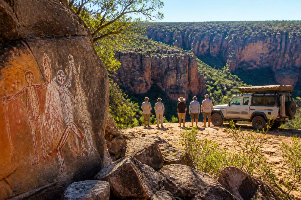 A small group of mature travellers standing beside a purpose-designed 4WD vehicle, looking out over a vast gorge with ancient rock art in the foreground, capturing the scale and remote beauty of the Kimberley