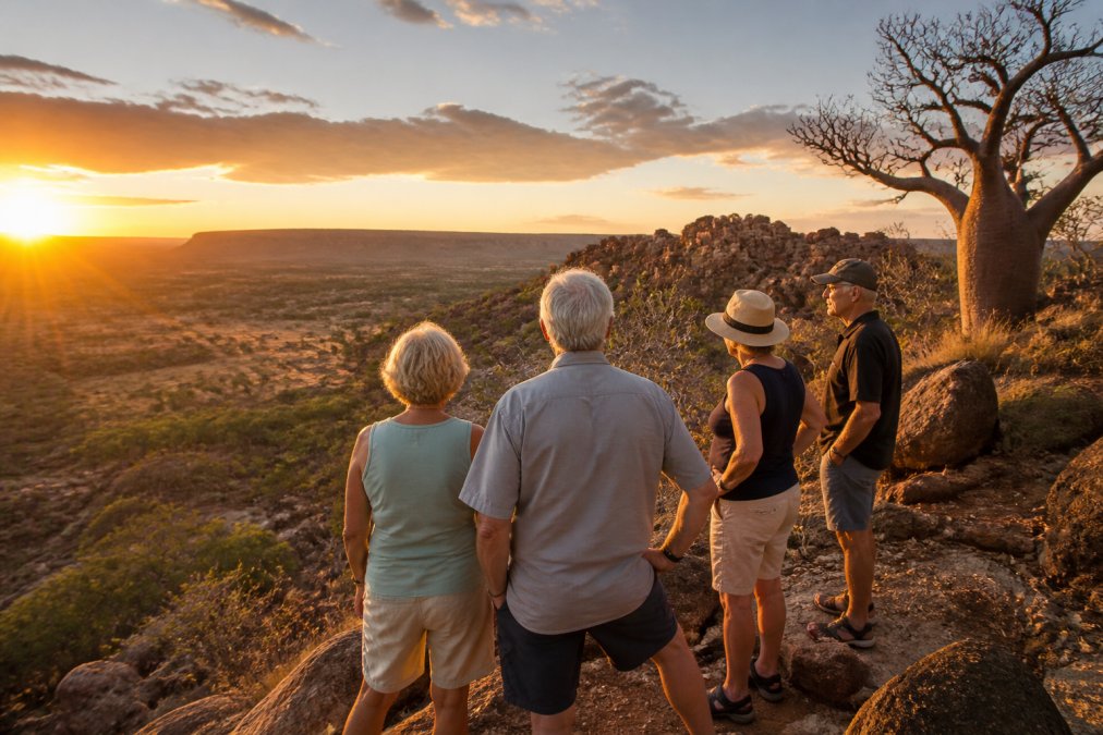 A small group of mature travellers standing at a scenic lookout overlooking the rugged Kimberley landscape at sunset, with boab trees and red rock formations in the background