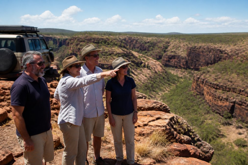 Small group of mature travellers standing together at a scenic gorge overlook in the Kimberley, smiling and pointing at the landscape, with a purpose-designed 4WD vehicle visible in the background
