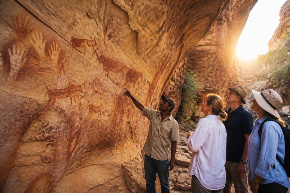 Mature travellers standing before ancient Aboriginal rock art in a remote gorge, guide gesturing to ochre hand stencils and animal figures on sandstone wall, golden afternoon light streaming through canyon opening