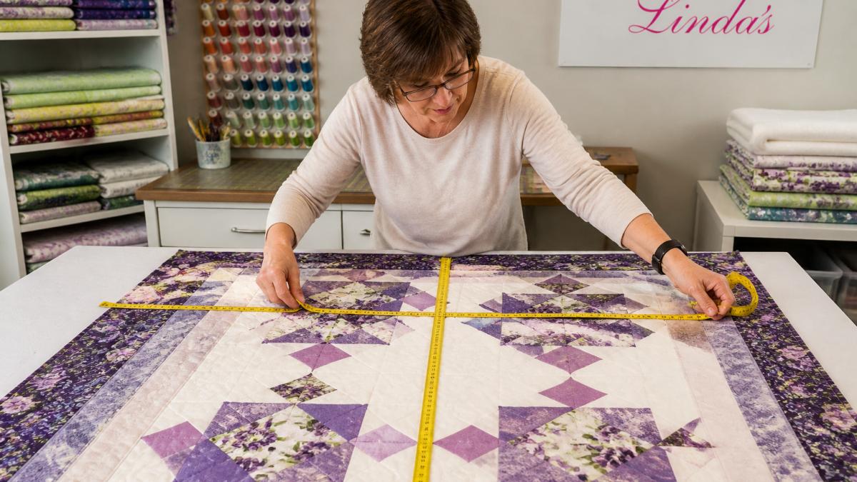 Quilter measuring a quilt top with a measuring tape on a design table, showing width and length measurements with fabric pieces visible