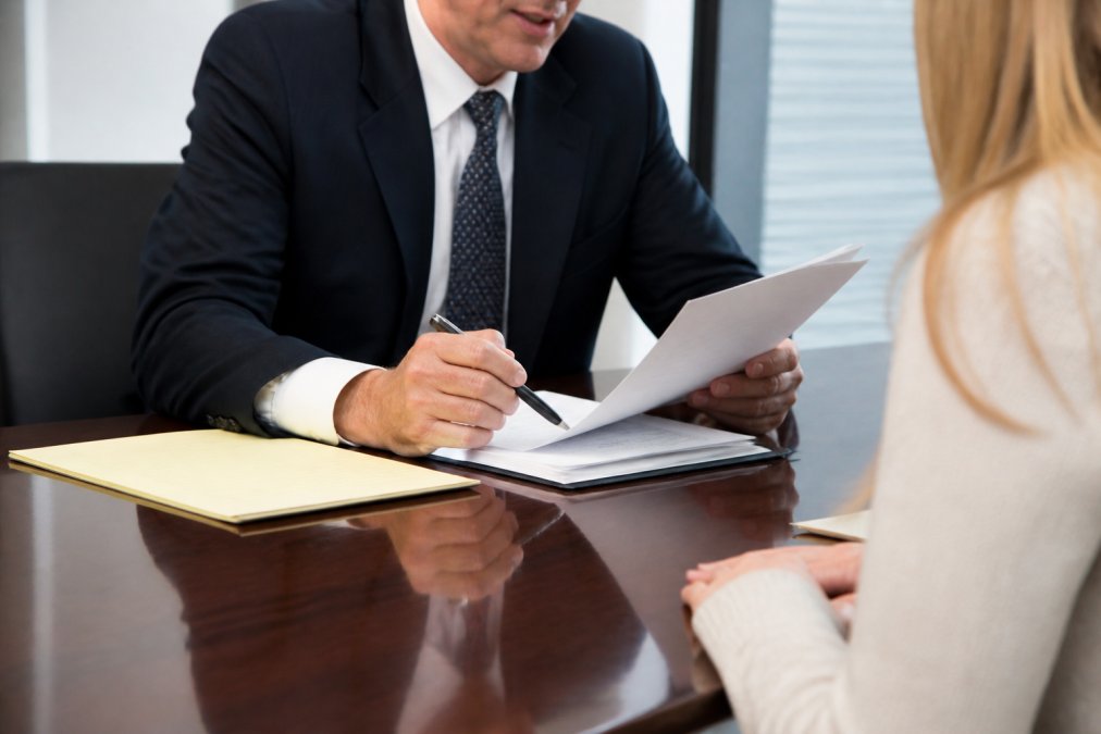 A professional attorney in a dark suit meeting with a client at a polished conference table, reviewing documents under bright office lighting