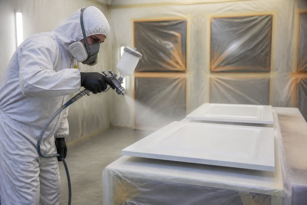 Professional painter in full protective gear and respirator spray-painting kitchen cupboard doors in a controlled spray booth, with masking tape and protective sheeting visible, demonstrating proper technique and equipment setup