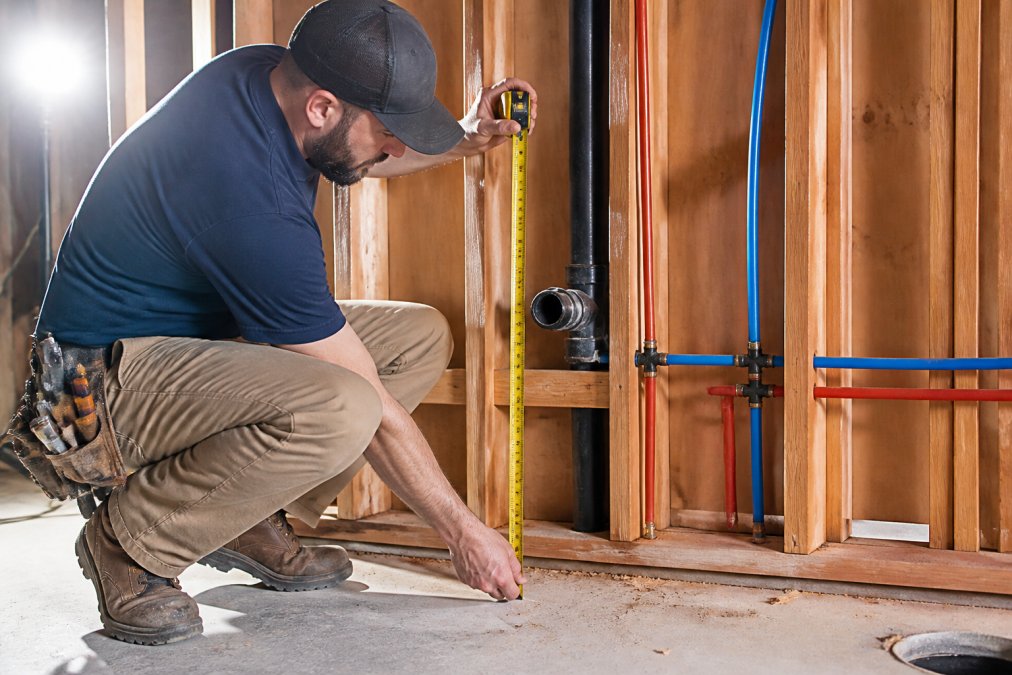 A professional plumber in work clothes measuring the distance from the finished floor to the drain stub-out with a tape measure in a kitchen under construction, exposed wall studs and PEX supply lines visible, bright work lights casting shadows