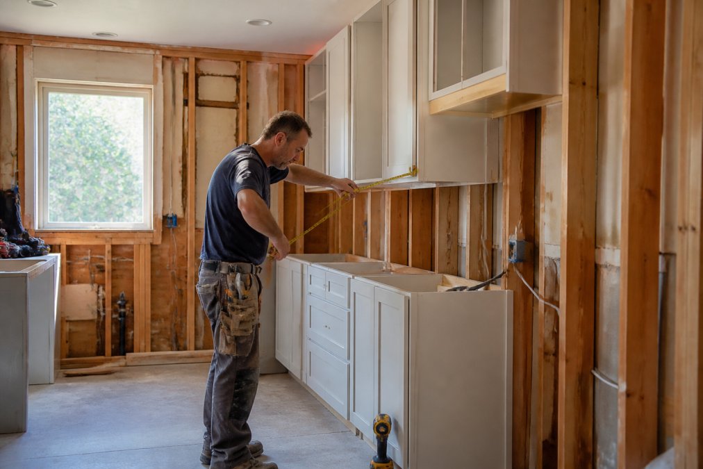 Professional contractor installing custom cabinetry in partially renovated kitchen with exposed studs, measuring tape in hand, and natural light from window