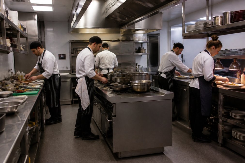 Professional commercial kitchen with clearly defined prep stations on the left, cooking range in the centre, plating pass on the right, with staff working efficiently at their designated stations in a well-lit space