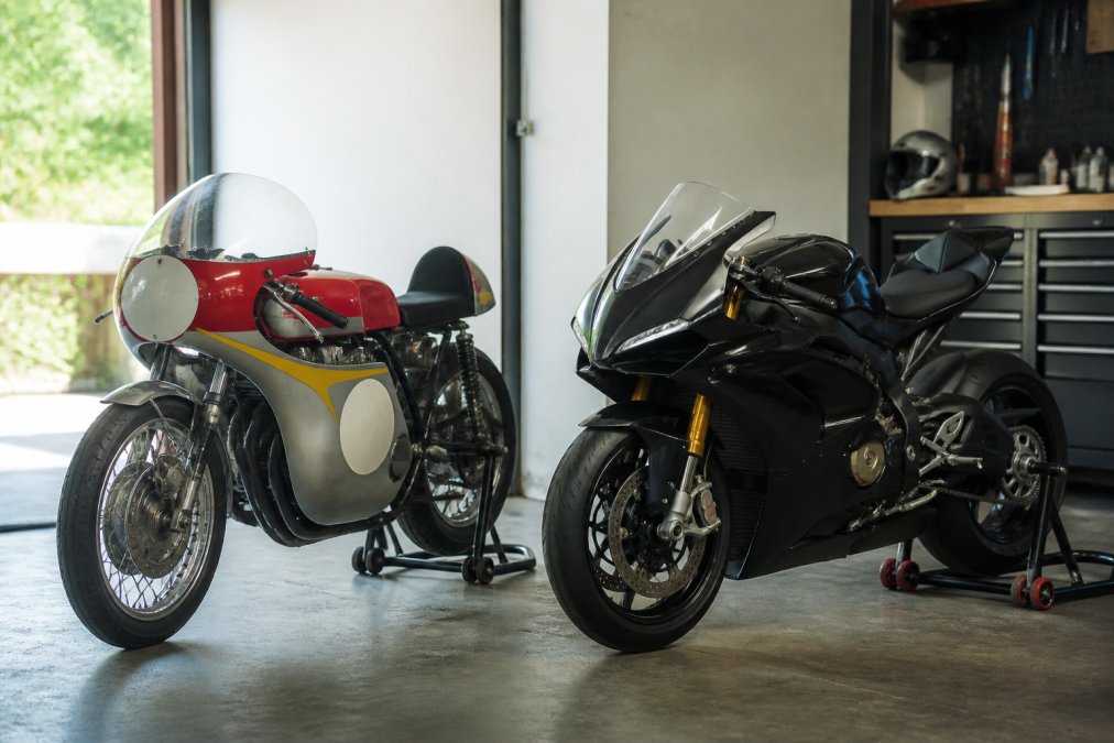 Een klassieke racemotor met zichtbare carburateurs en een moderne superbike met digitale cockpit naast elkaar in een goed verlichte garage, beide in perfecte staat
