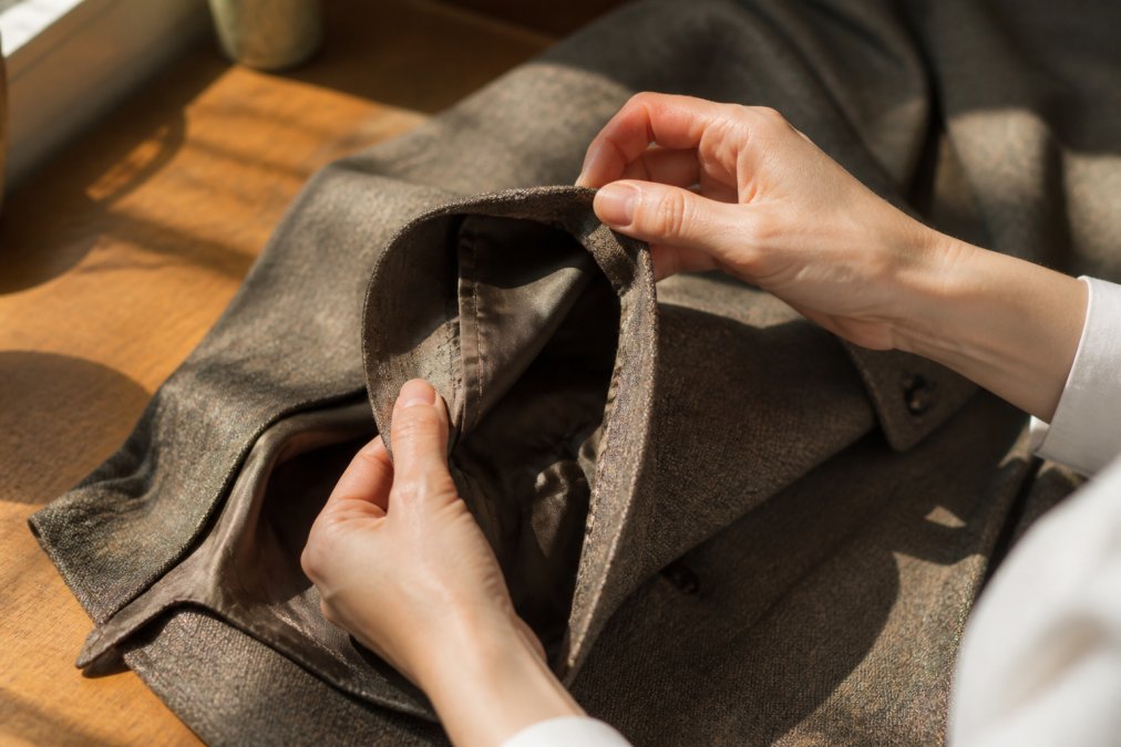 Nahaufnahme von Händen, die die Innennaht und den Stoff eines Vintage-Blazers untersuchen, warmes Tageslicht fällt durch ein Fenster auf den Tisch