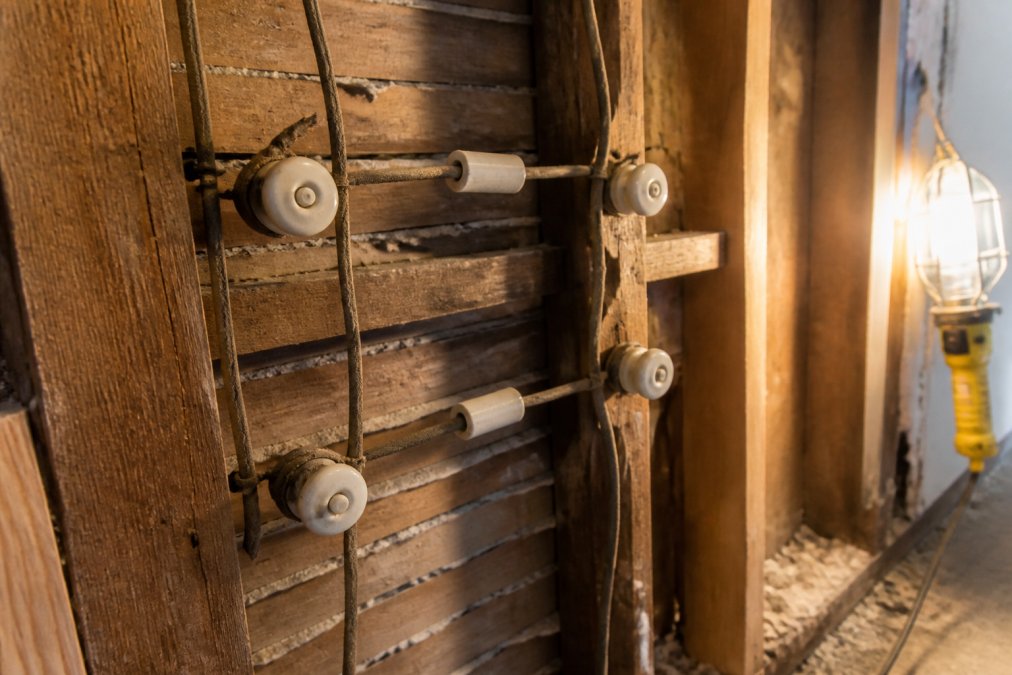 Close-up of old knob and tube wiring visible inside an open wall cavity, showing ceramic knobs, cloth-wrapped copper wires, and aged porcelain tubes under warm incidental lighting from a work lamp