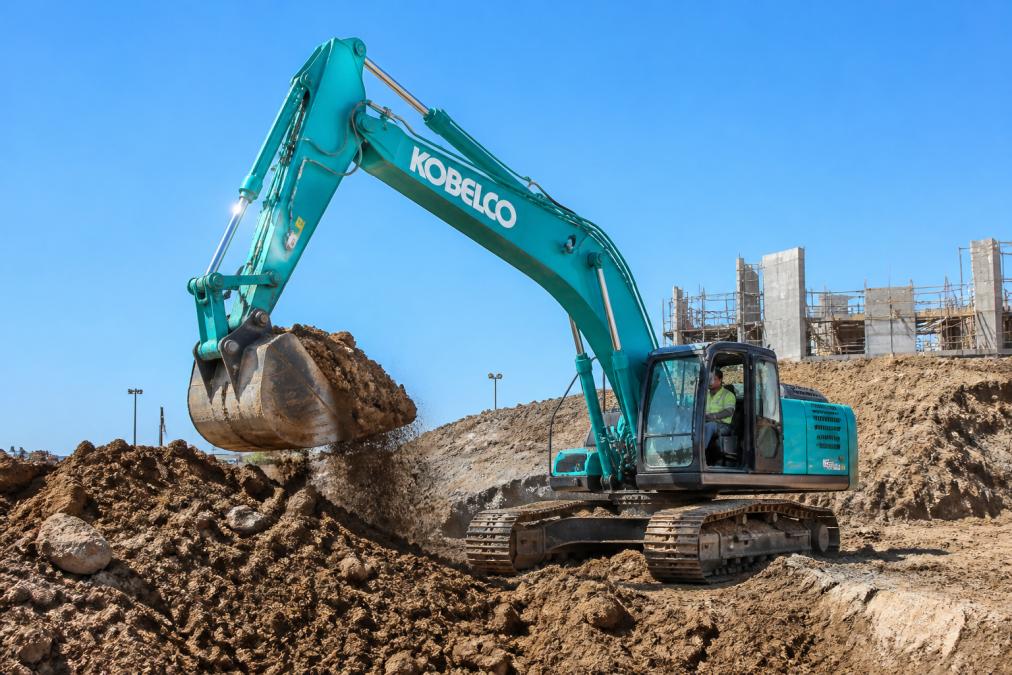 Kobelco hydraulic excavator moving dirt at a sunny construction site with an operator in the cab