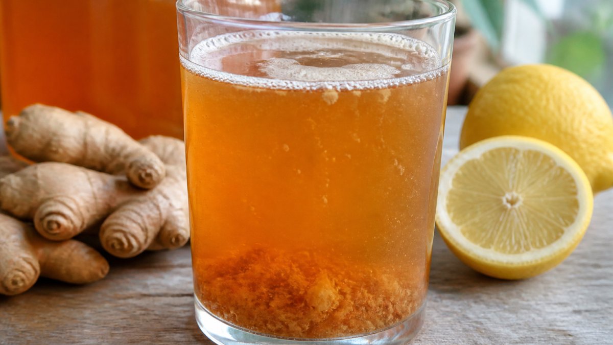 Close-up of a glass of raw kombucha with visible sediment and SCOBY particles, showing the fermented beverage in natural light with fresh ginger and lemon nearby