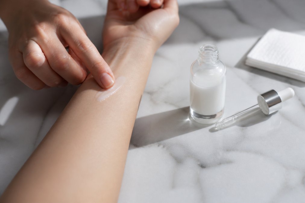 Close-up of hands applying a small amount of serum to the inner arm for patch testing, with a clear glass bottle and white cosmetic product visible on a clean marble surface in natural daylight