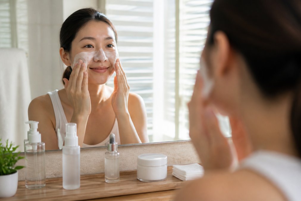 Woman applying Korean skincare products in sequence on her face, cleansing, applying toner mist, patting in serum, and finishing with moisturizer, in a bright, modern bathroom with natural morning light