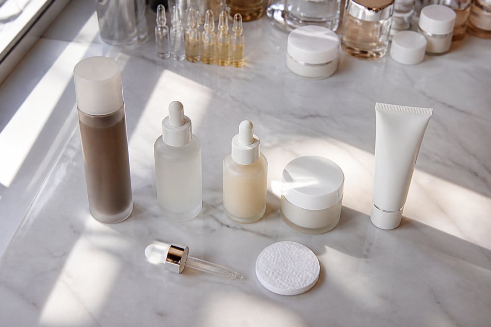 Flat lay of multiple Korean skincare products arranged in application order on a white marble surface, toner, essence, serum, cream, and sunscreen bottles neatly lined up with a dropper and cotton pad nearby, soft natural light from the left