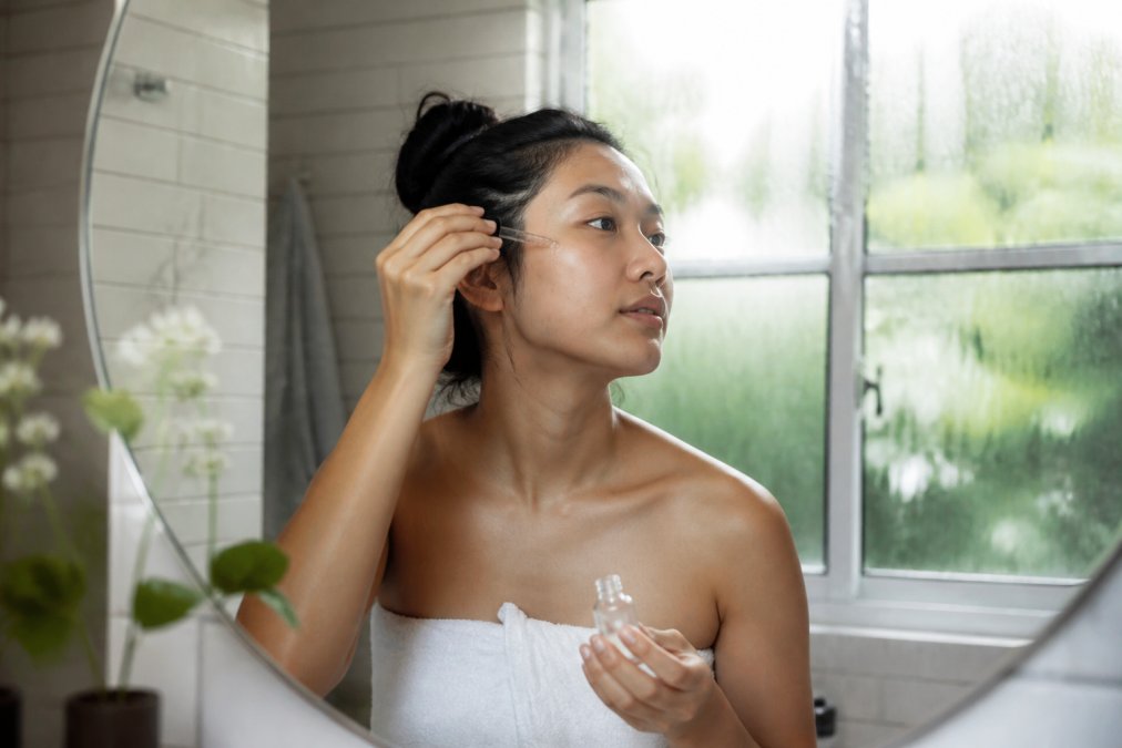 Person applying lightweight hydrating serum to clean face in bright, humid bathroom with condensation visible on mirror and natural light streaming through window