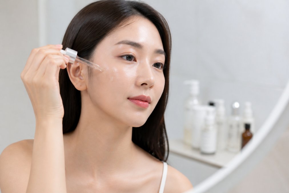 Close-up of a woman's hands applying a drop of serum to her cheek in a bright, minimal bathroom setting, with skincare bottles softly blurred in the background