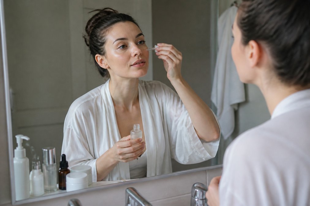 A woman in her thirties applying serum to her face at a bathroom mirror in soft morning light, skincare bottles arranged on the counter beside her