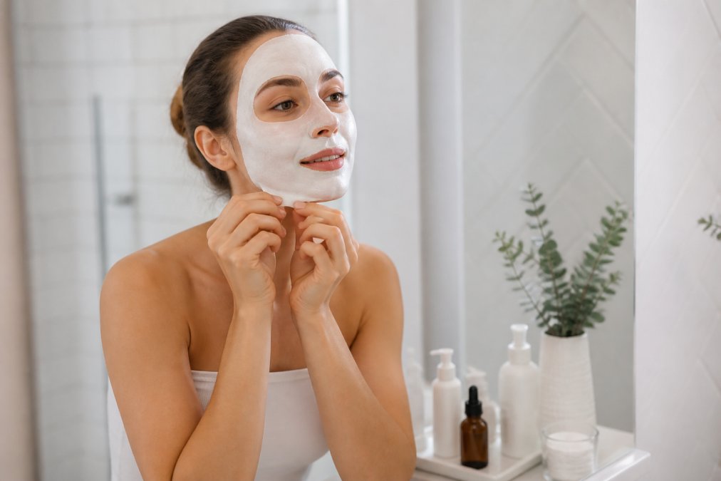 A woman applying a white peel-off face mask to her clean skin at home in a bright, minimalist bathroom, looking relaxed and focused, with a soft overhead light and a small collection of skincare products on the counter beside her