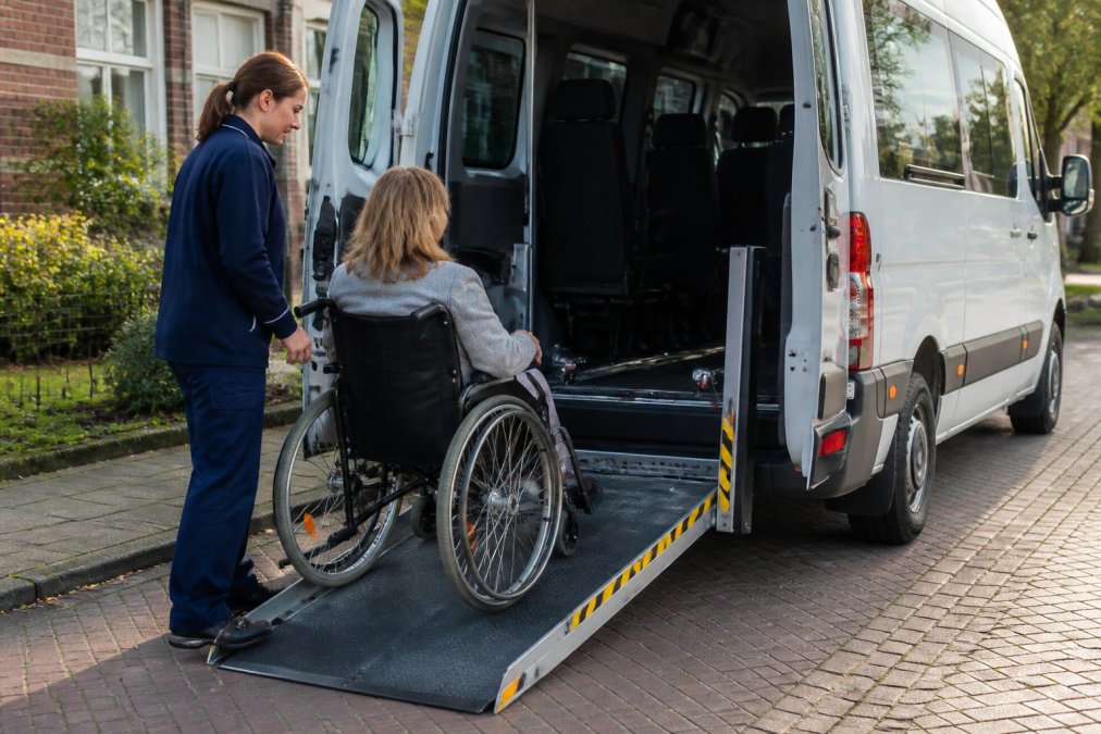 Close-up van een uitgeschoven oprijplaat aan de achterzijde van een witte rolstoeltaxibus, met een zorgverlener in uniform die naast de plaat staat klaar om een passagier te begeleiden, op een rustige straat op een zonnige dag in Nederland